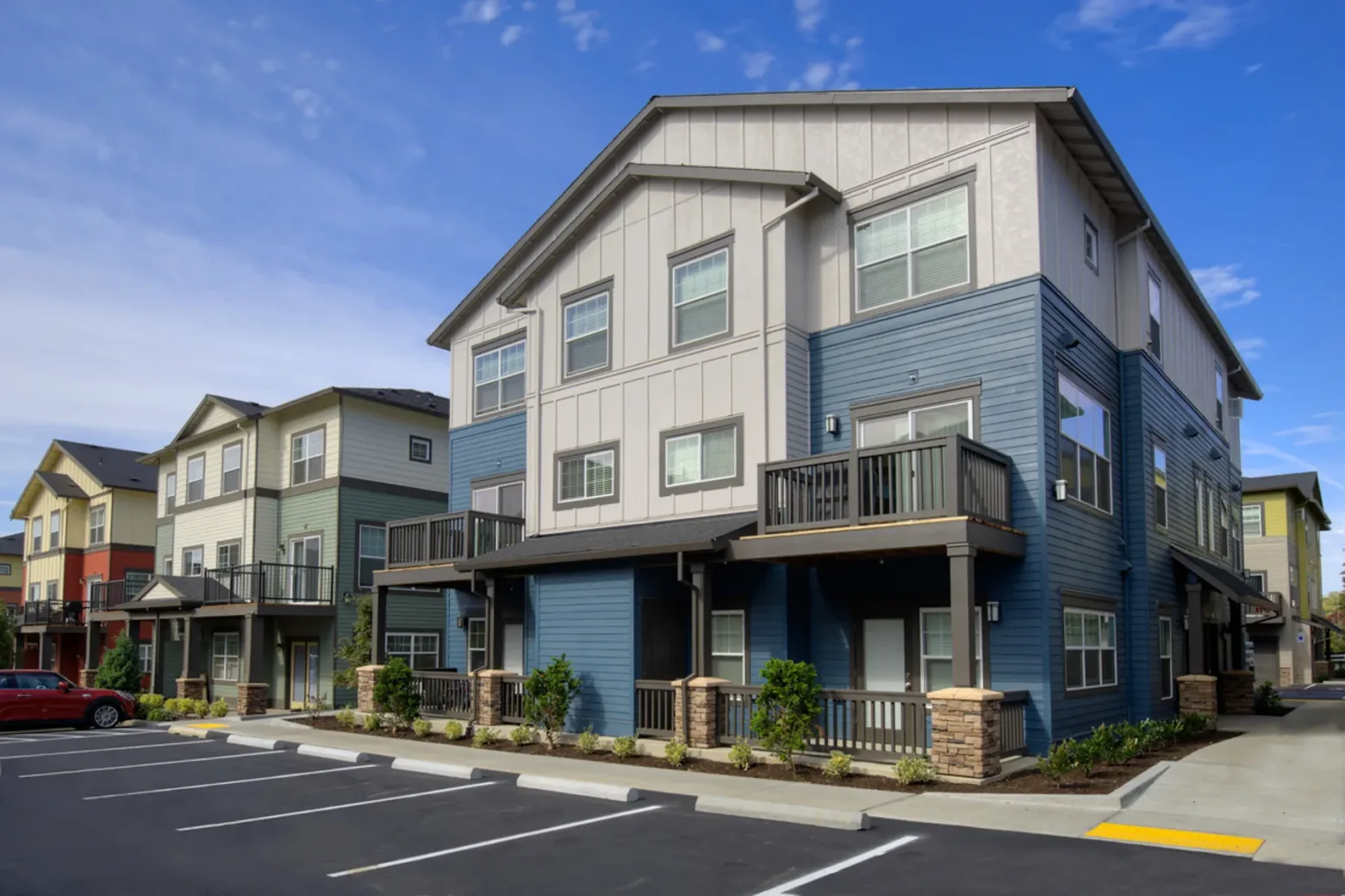 A modern apartment complex with multiple two-story buildings, featuring blue and beige siding, balconies, and landscaped surroundings, set under a clear blue sky.