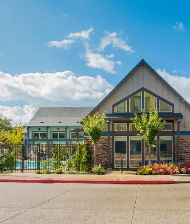 A modern apartment complex with a prominent clubhouse featuring large windows, landscaped gardens, and a fenced swimming pool area, under a bright blue sky with scattered clouds.