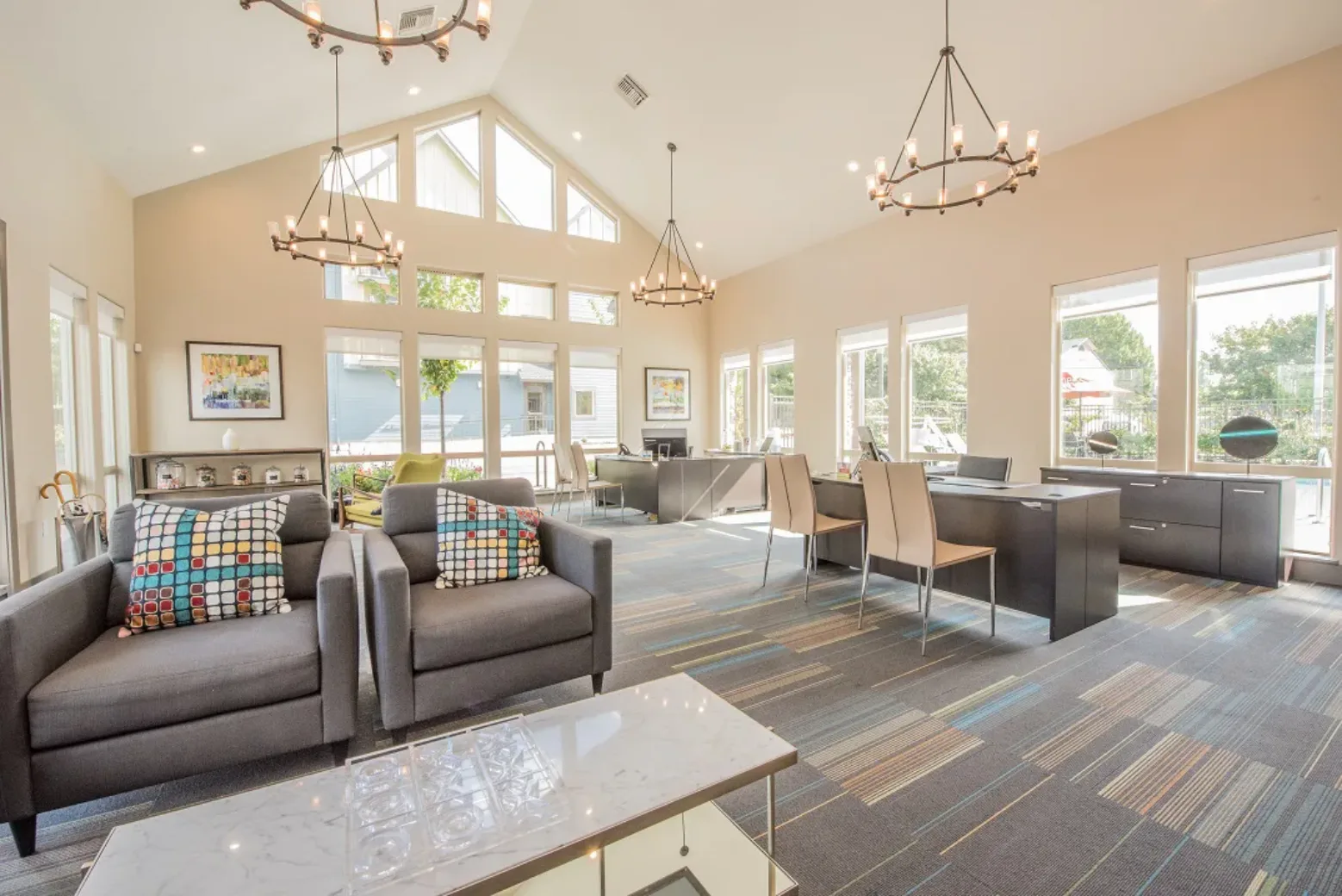 Bright and spacious office lounge with large windows, grey sofas, colorful cushions, chandeliers, and modern desks.