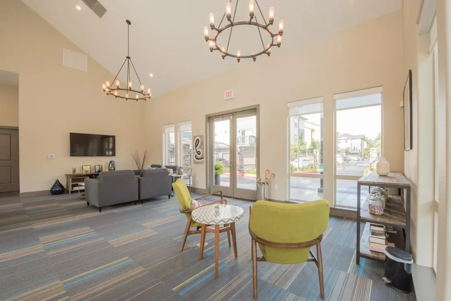 A spacious apartment complex lounge featuring modern furniture including two gray sofas and a pair of green chairs around a small table. Large windows allow natural light to fill the room, and two large chandeliers hang from the ceiling. The flooring is a carpet with a striped pattern, and a flat-screen TV is mounted on the wall. A glass door provides access to an outdoor area.