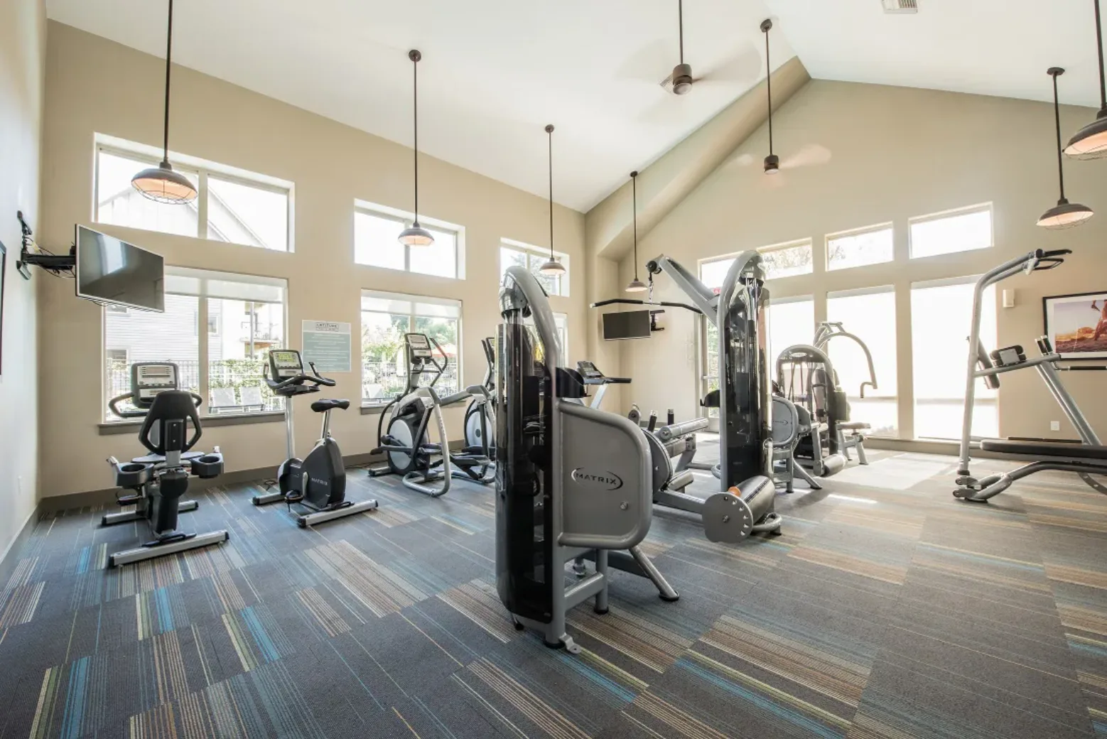 A bright gym room with various exercise machines including treadmills, stationary bikes, and weight lifting equipment. There are large windows allowing natural light and a TV mounted on the wall.