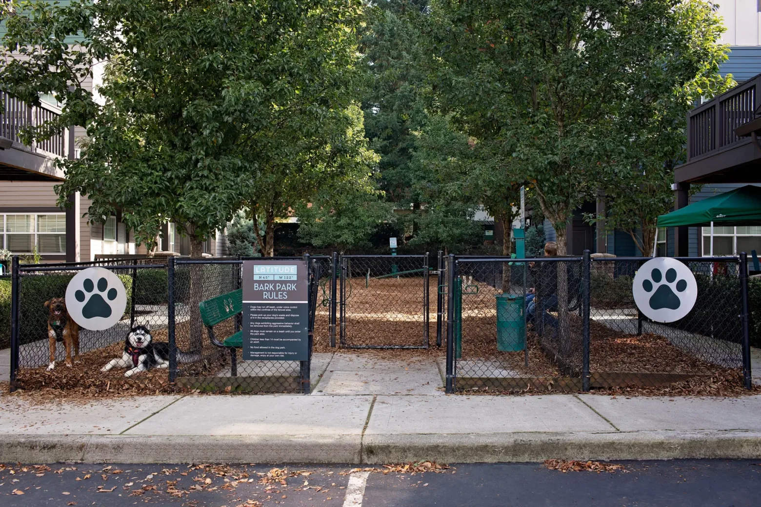 A fenced dog park area in an apartment complex, featuring two dogs inside, with trees and a sign displaying park rules. The fence is decorated with large paw print signs.