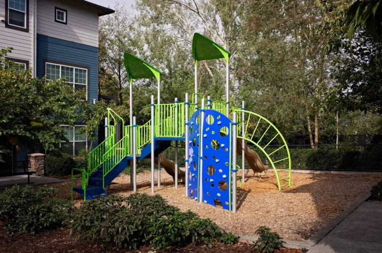 Outdoor playground with vibrant green and blue equipment surrounded by trees and part of an apartment building visible in the background.