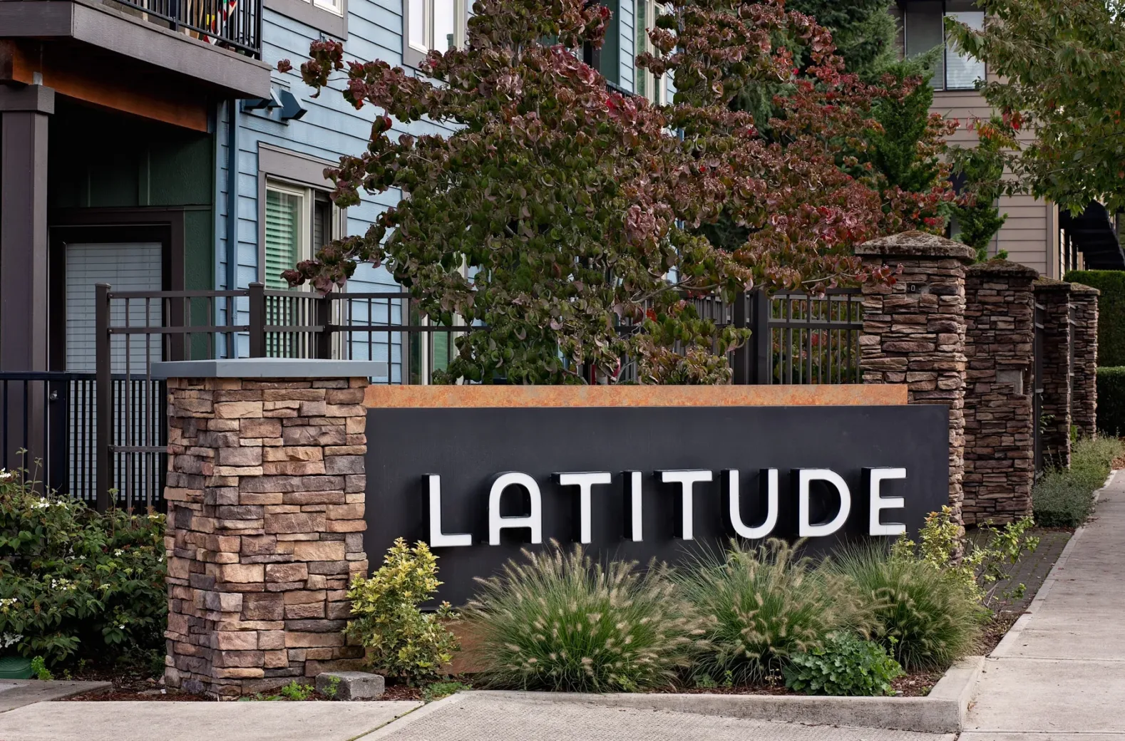 Sign reading 'LATITUDE' on a stone pillar at an apartment complex entrance, surrounded by landscaping with bushes and trees.