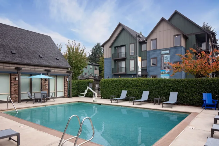 An apartment complex with a central outdoor swimming pool surrounded by lounge chairs and tables. The buildings feature a mix of blue and gray siding with pitched roofs, set against a backdrop of trees and a clear sky.