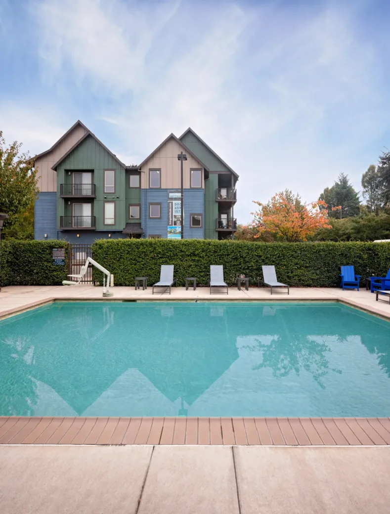 Outdoor swimming pool with surrounding lounge chairs in front of a multi-story apartment building with green, blue, and beige paneling.