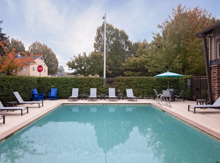 Outdoor swimming pool with lounge chairs and tables under umbrellas, surrounded by greenery and adjacent to a building.
