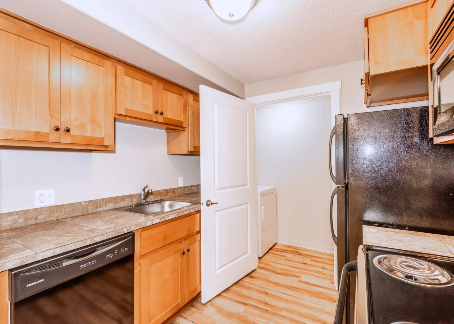 A kitchen with light wood cabinets, a refrigerator, dishwasher, and an electric stove. There is a stainless steel sink and tiled countertop. A door is open, revealing a small laundry area with a washer and dryer.