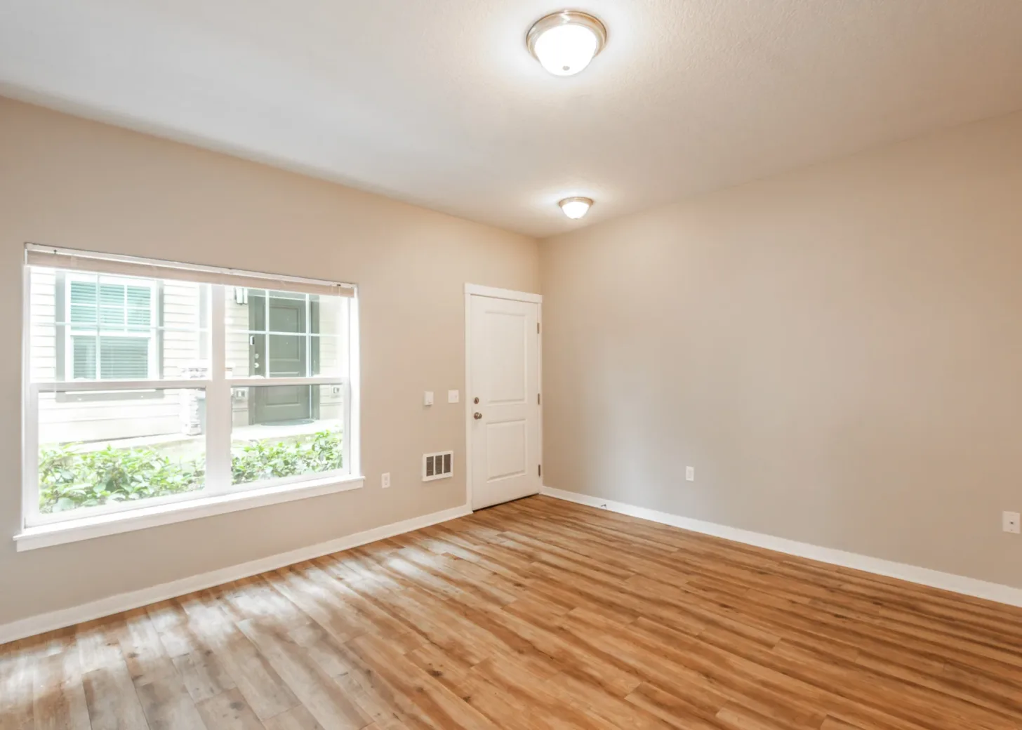 Interior of an empty apartment room with wooden flooring, beige walls, and a large window allowing natural light from outside.