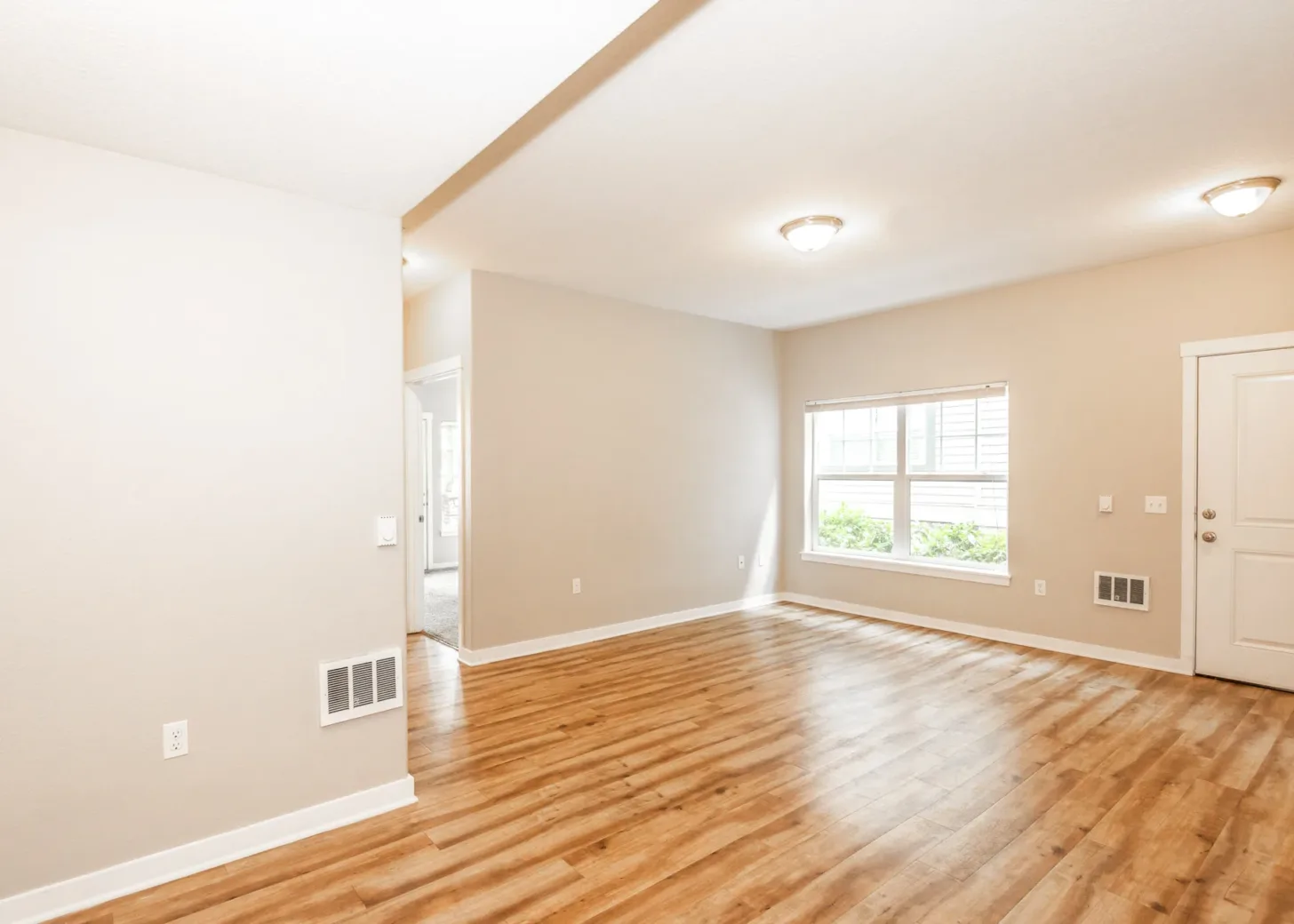 A spacious apartment interior with light wooden floors, beige walls, and ceiling lights, featuring a window and door leading to another room.