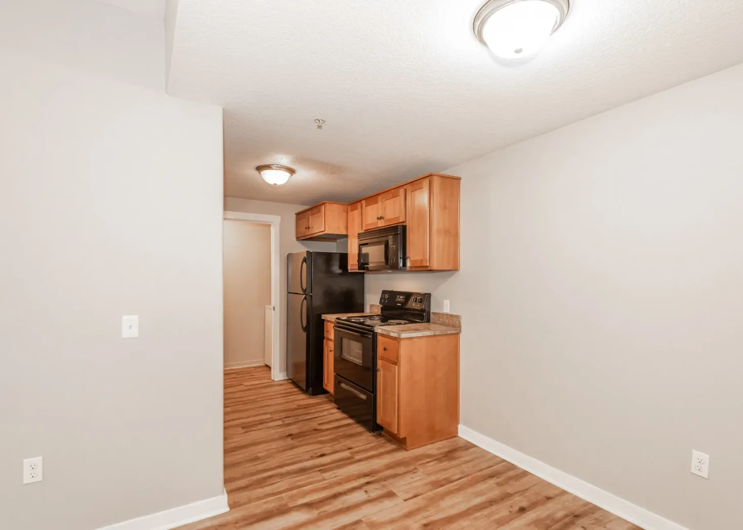 Kitchen area in an apartment featuring light brown wooden cabinets, black appliances, and hardwood flooring.