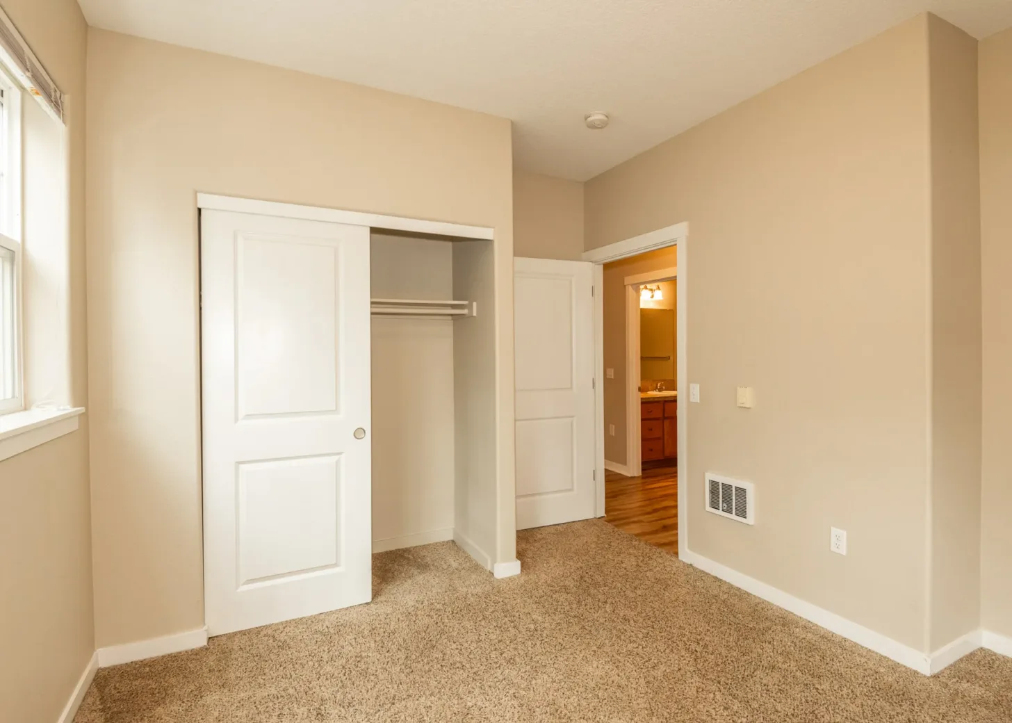 Interior view of an apartment bedroom featuring beige carpets, a closet with sliding doors, and an open door leading to a bathroom.