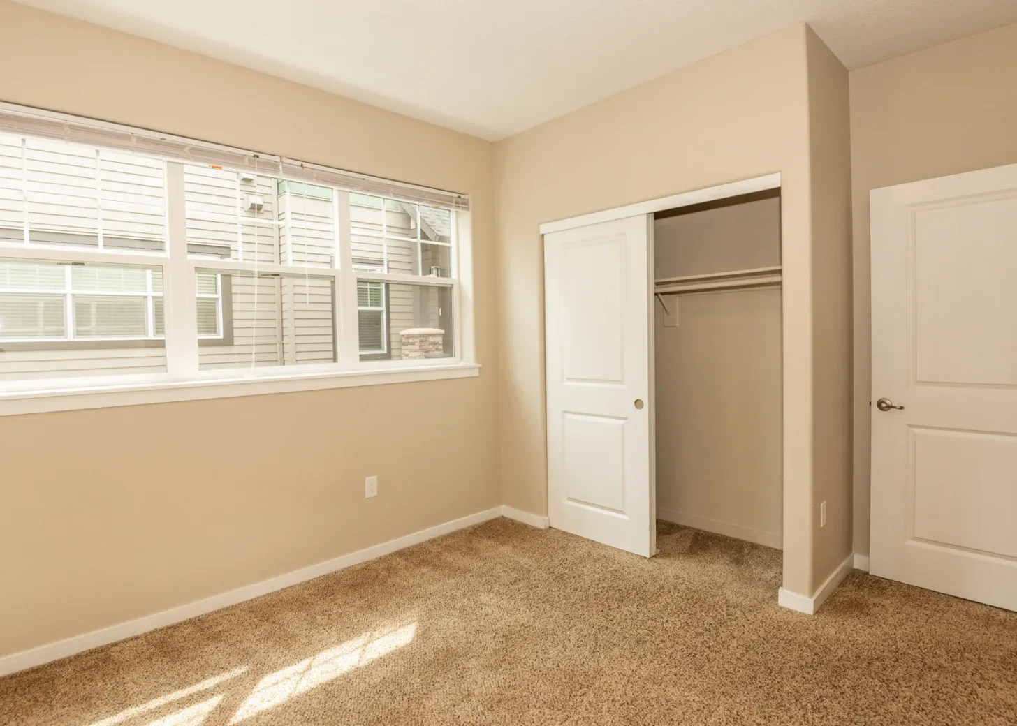 Interior view of an empty apartment room with beige walls, carpeted floor, a large window, and a closet with sliding doors.