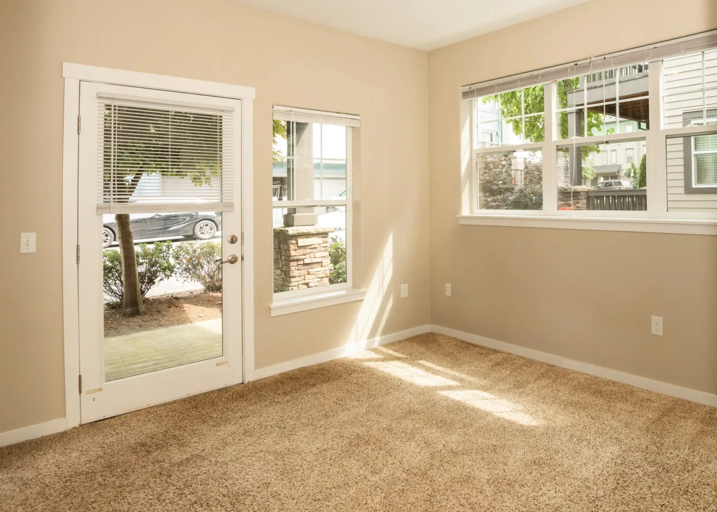 Sunlit corner of an apartment with beige carpet, a glass door, and windows overlooking a residential area.