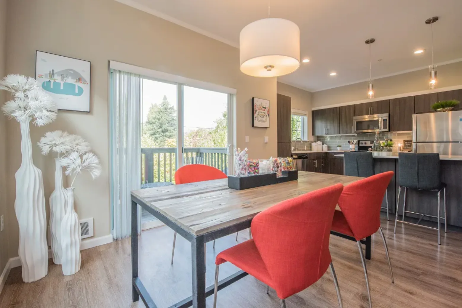 Modern apartment dining area with red chairs around a wooden table, adjacent to a kitchen with stainless steel appliances and a view of trees through a sliding glass door.
