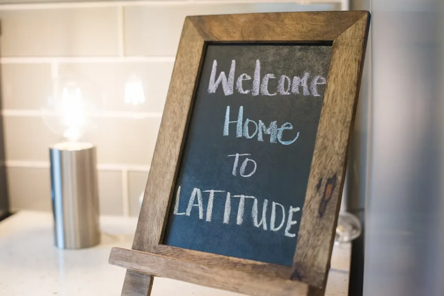 A small chalkboard in an apartment reads 'Welcome Home to Latitude,' placed on a kitchen countertop with a modern lamp in the background.