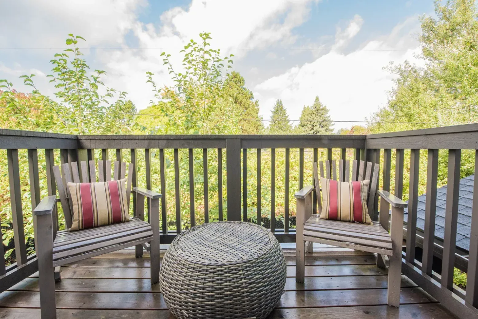 Balcony of an apartment with two wooden chairs and a woven round table, overlooking lush greenery.