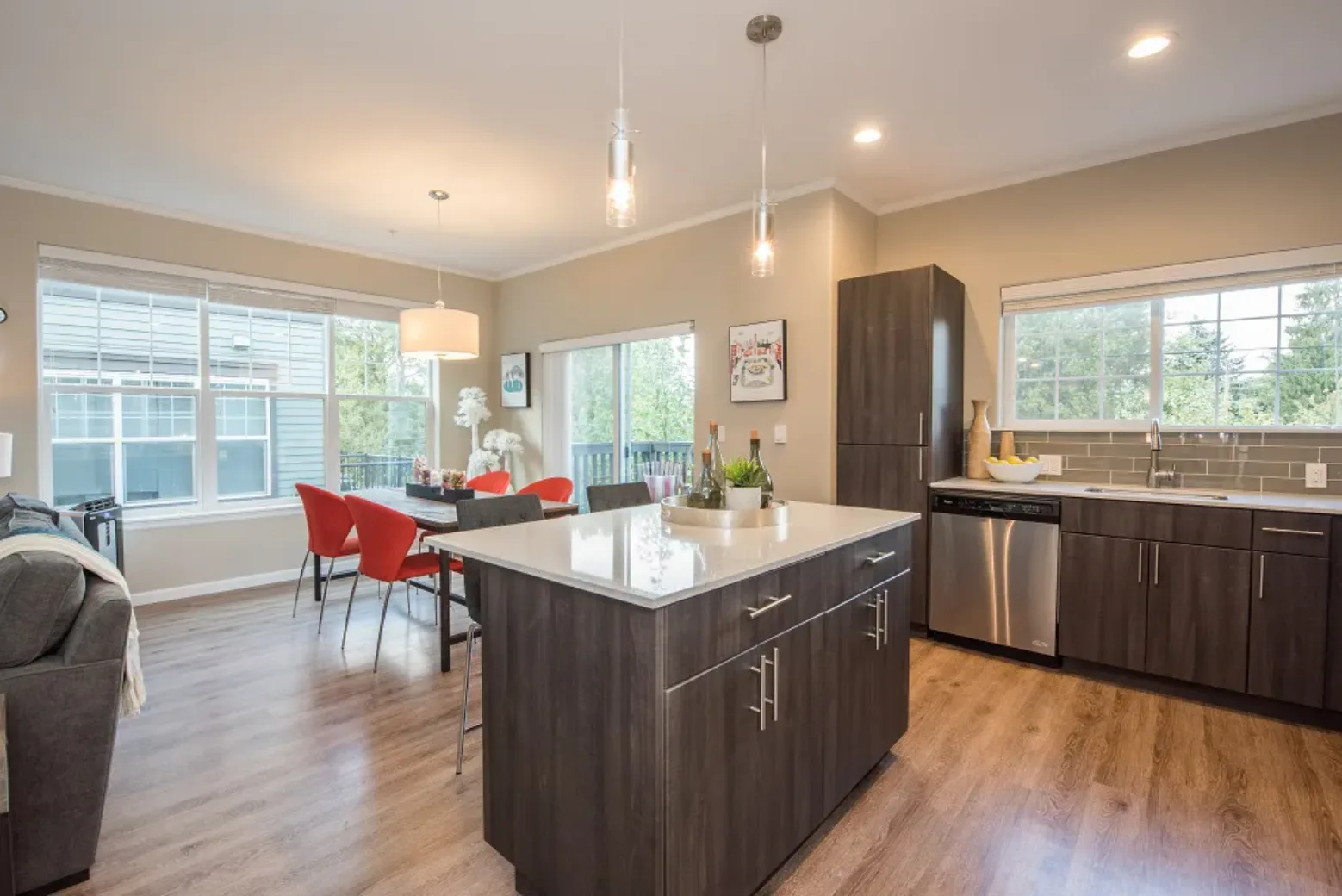 Modern apartment interior featuring an open-plan kitchen and dining area with wooden flooring, dark cabinetry, an island, and large windows providing natural light.