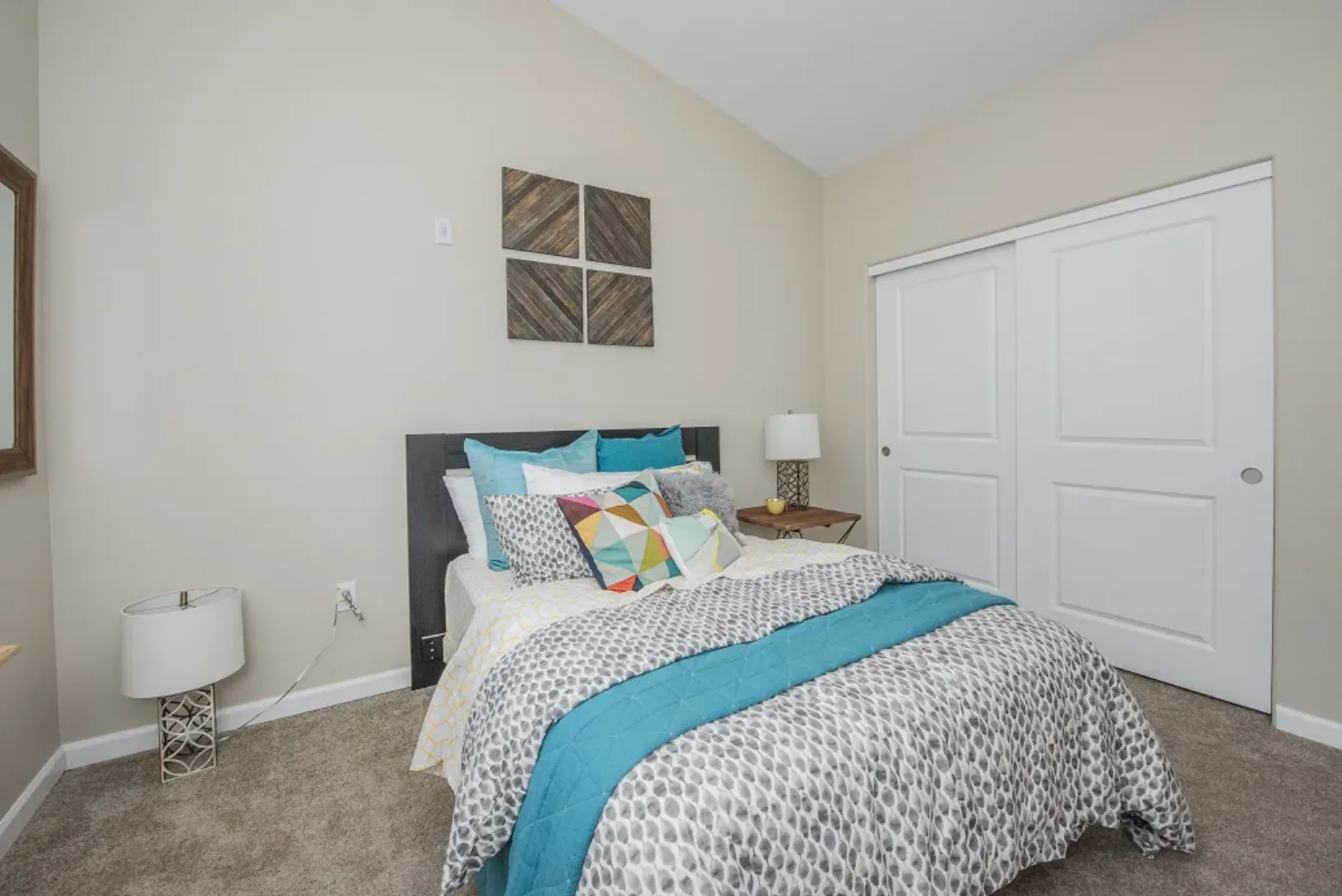 Bedroom in an apartment with a bed featuring colorful pillows and a blue comforter, a side table with a lamp, and sliding closet doors.