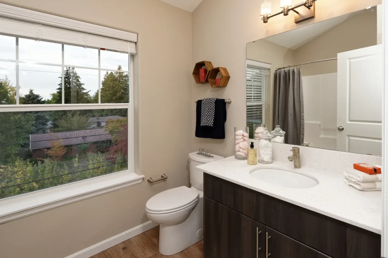 Bathroom in an apartment featuring a large window with an outdoor view, a toilet, a sink with a countertop, and wall-mounted shelves and lighting.