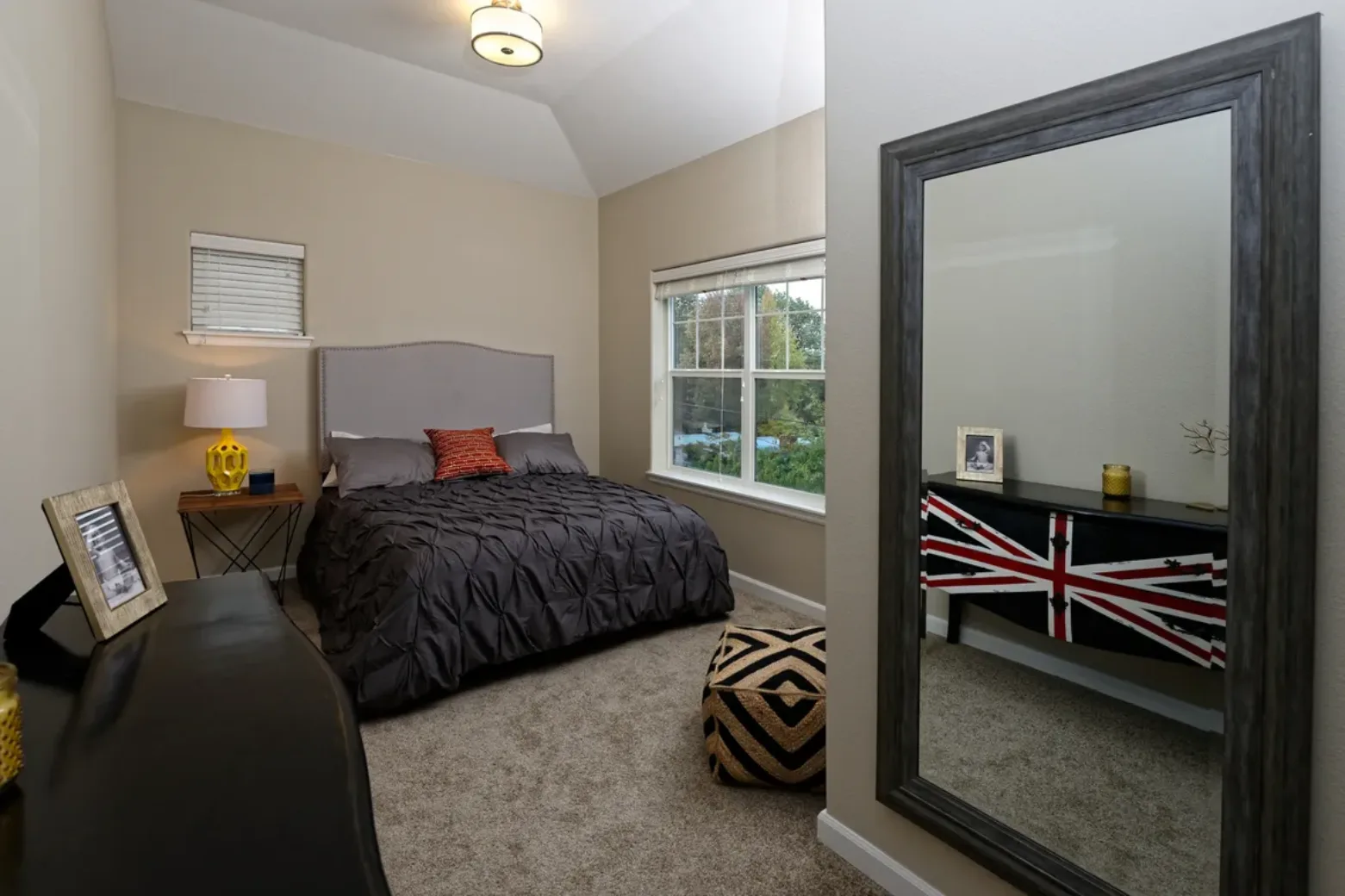 Apartment bedroom with a gray bed, large mirror, and a dresser featuring a Union Jack design, alongside a window with a view of greenery.