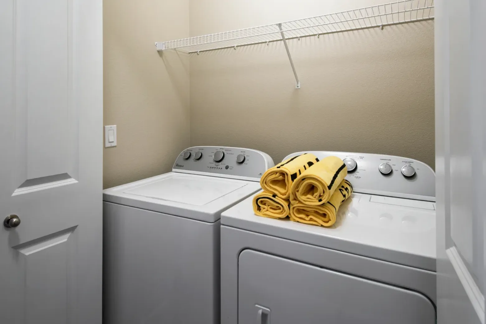Apartment laundry room with a washing machine and dryer, featuring a stack of folded yellow towels on top and a wire shelf above.