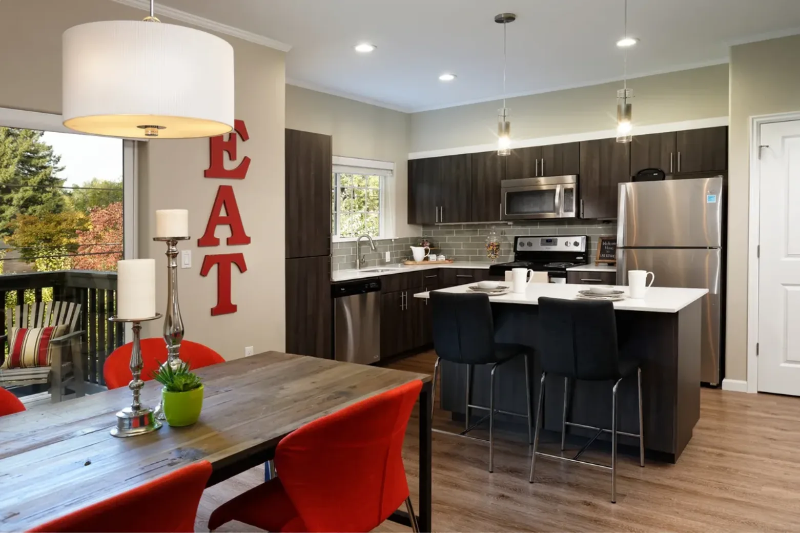 Modern kitchen and dining area in an apartment, featuring dark wood cabinets, stainless steel appliances, a small kitchen island with seating, and a dining table with red chairs beneath a large hanging lamp.