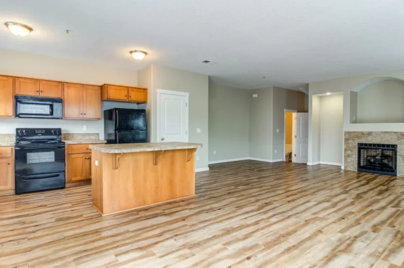 An open floor plan kitchen and living area with wood laminate flooring, featuring wooden cabinetry, a black stove, and refrigerator, a small kitchen island, and a fireplace set into one wall.